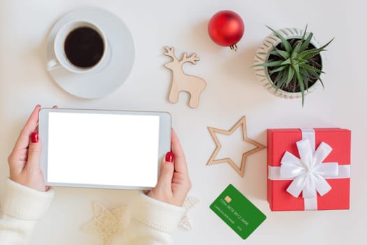 On a white table a woman's hands with a digital tablet, next to a green bank credit card, a red gift box with a bow, a cup of coffee and a green succulen in a pot, Christmas decor. Mock up.