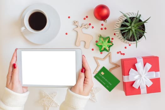 The hands of a woman are holding a digital tablet, a blank credit card, a red gift box , a cup of coffee and a succulent plant , Christmas decor and toys are lying next to the white table. Mock up