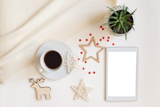 A digital tablet with a blank screen, lies on a white table surrounded by flat wooden Christmas decorations, a deer, stars, snowflakes, a white cup with coffee.Top view. Mock up