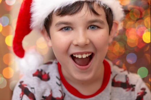 Close-up portrait of a handsome joyful European boy in a red Santa Claus cap against the background of bokeh of New Year garlands.