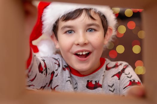 A surprised, happy, handsome European boy in a New Year's jumper and Santa Claus hat takes a present out of the box, holding out his hands to the present, on the background of bokeh of lights garlands