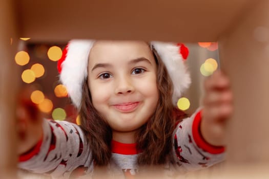 A happy girl in a New Year's jumper and Santa Claus hat takes a present out of the box, holding out his hands to the present, on the background of bokeh of lights garlands