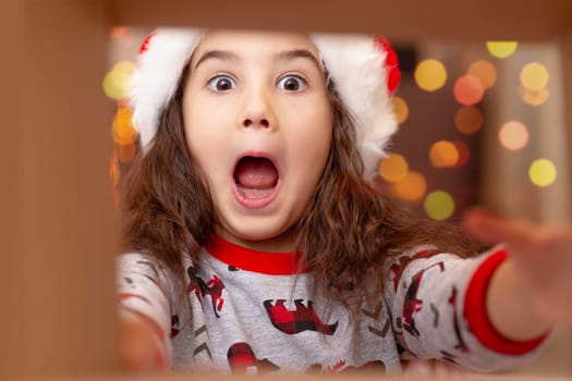 Close-up portrait. A joyful, surprised, beautiful girl in a New Year's jumper and a Santa Claus hat takes a gift out of the box, stretching out her hands to the gift, view from the inside.