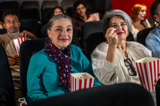 Two senior women eating popcorn and watching a movie in the cinema, enjoying their leisure time