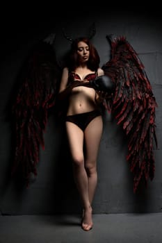 Full body of seductive female with horns and black wings looking at camera on black background with skull