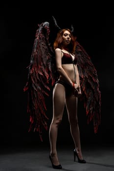 Full body of seductive female in lingerie and wings with horns looking at camera on black background in dim studio
