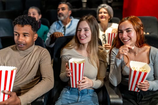 Happy multi ethnic friends watching film together and eating popcorn, having fun at the movie theatre