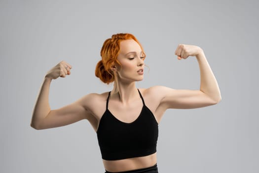 Self assured young fit female model with ginger hair in black sportswear showing biceps while standing against gray background