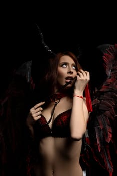 Seductive female in underwear with horns and wings looking up while touching neck collar on black background in dark studio