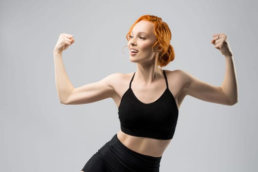 Self assured young fit female model with ginger hair in black sportswear smiling and showing biceps while standing against gray background and looking away