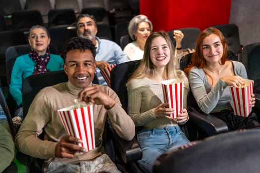 Multi ethnic group of friends enjoying a film at the movie theater, sharing laughter and munching on popcorn in a cozy auditorium setting