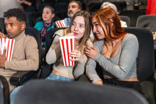 Two friends are whispering and eating popcorn while watching a movie at the cinema, surrounded by other people