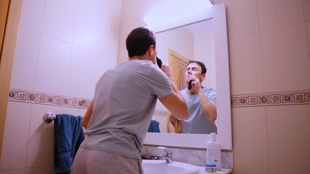 Man using a small electric trimmer to remove his nose hair while looking in the bathroom mirror during his morning routine