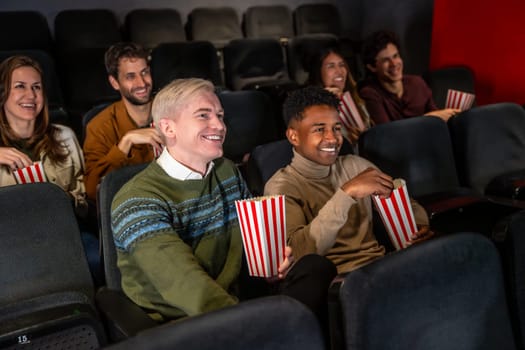 Diverse group of friends laughing and eating popcorn while watching a movie at the cinema
