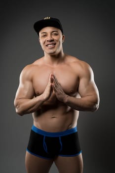 Full body of sexy shirtless trainer in underpants and cap showing muscles and looking at camera on gray background in studio
