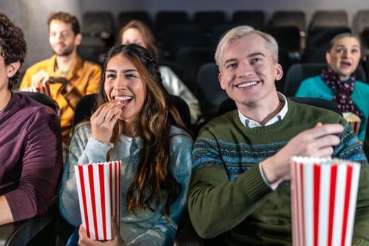 Young couple enjoying a movie at the cinema, eating popcorn and smiling, surrounded by other people