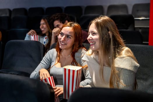Two young women enjoying popcorn and smiling while watching a movie together in a cozy cinema, sharing laughter and a fun evening