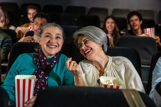Two cheerful senior women laughing and eating popcorn while watching a movie at the cinema, sharing a joyful moment together