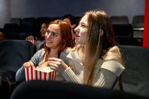 Two young women smiling and enjoying popcorn while watching a movie together in a cozy cinema, sharing laughter and a fun evening