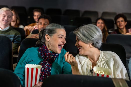 Two cheerful senior women sharing popcorn and laughing together while watching a movie at the cinema, enjoying a fun night out