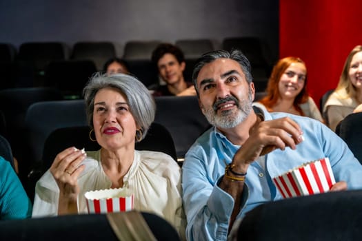 Senior couple watching a movie and eating popcorn, enjoying a night out at the cinema with other people