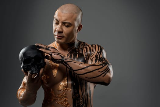 Handsome sexy male with flowing paint stains on fit naked torso touching black skull on gray background in studio