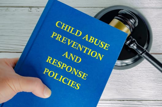 Hand holding Child Abuse Prevention and Response Policies book with gavel background.