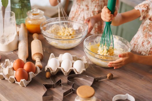 Children whisk dough in glass bowls surrounded by fresh eggs, baking tools, and flour on a wooden table.