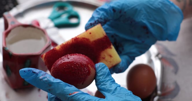 Hands wearing blue gloves carefully apply red dye on a brown egg, highlighting the joyful activity of coloring eggs for Easter celebrations, adding creativity to the holiday spirit.