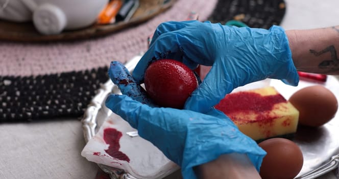 Hands wearing blue gloves hold a red Easter egg being painted as part of a coloring activity. Surrounding items include brown eggs and dye, hinting at the joy of spring celebrations.