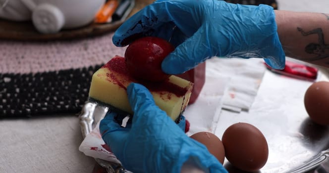Hands, protected with blue gloves, carefully coat a red Easter egg using a sponge while decorating eggs for the holiday. This creative activity brings family together during springtime festivities.