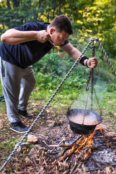 Young camper tasting food from a cauldron hanging over a campfire, enjoying a wilderness cooking experience