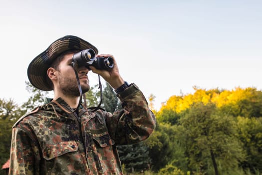 Soldier using binoculars while observing enemy positions in forest during military operation