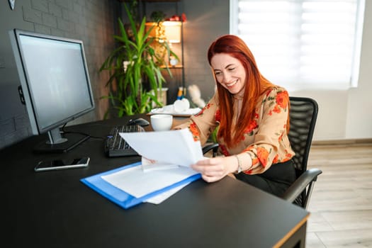 Smiling red-haired woman arranges bills on her desk.