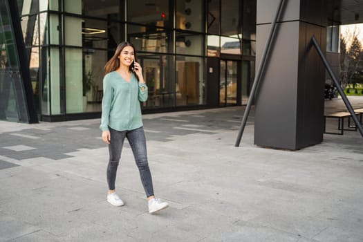 Smiling young brunette girl walks in outdoors and makes a phone call.