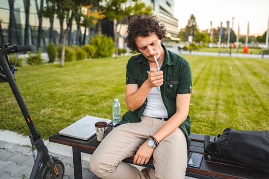 Man with curly hair sits on a park bench, lights a cigar and rests after riding an electric scooter