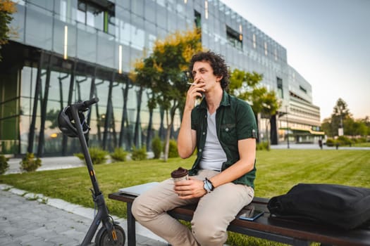 Man with curly hair sits on a park bench, smokes a cigar and rests after riding an electric scooter.