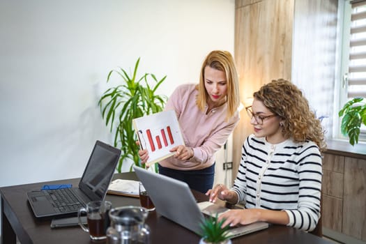 Two female colleagues are working from home, one is sitting at a desk using a laptop, while the other is standing next to her with a chart in hand, working together on a business report.