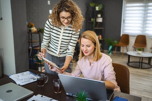 Two female colleagues are working from home, one is sitting at a desk using a laptop, while the other is standing next to her with a document in hand, working together on a business report.