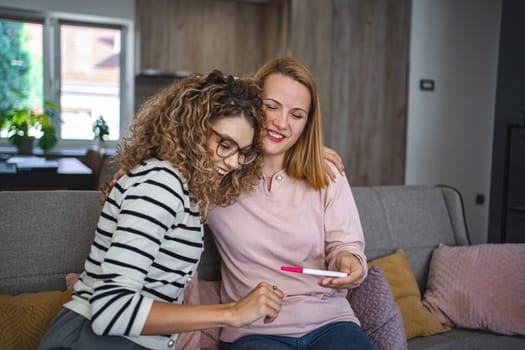 Two female friends are sitting on the couch in the living room and are excited after receiving the results of a pregnancy test