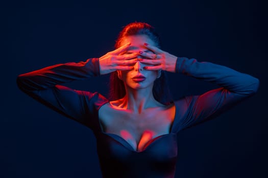 Studio portrait of a confident woman in bold red and blue lighting, covering eyes with hands, modern fashion pose.