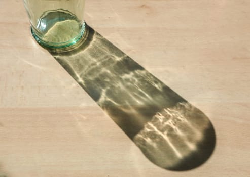 Empty glass bottle projecting an elongated shadow on a wooden surface, creating an interesting play of light and shadow