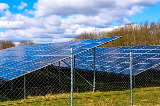 Rows of solar panels stand behind a wire mesh fence, converting sunlight into electricity under a bright blue sky