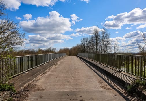 Empty narrow bridge with metal railing crossing a river in the countryside on a beautiful sunny spring day with some clouds