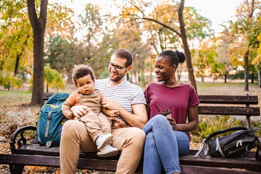 Mixed race couple is sitting on a park bench with their son.