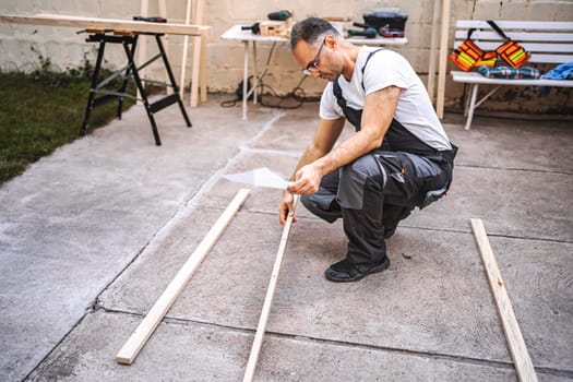 Mature male carpenter using electric screwdriver on a wooden plank outdoors.