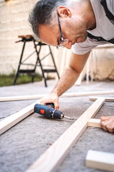 Mature male carpenter using electric screwdriver on a wooden plank outdoors.
