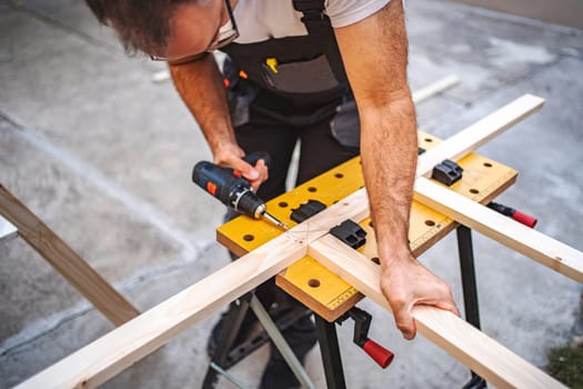 Mature male carpenter using electric screwdriver on a wooden plank outdoors.