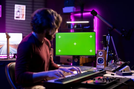Audio engineer playing electronic piano keyboard in home studio, recording tunes an watches computer with greenscreen after creating a melody on midi controller. Artist uses synthesizer notes.