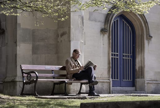 Windsor, UK - Apr 03, 2025 - An elderly man with glasses reading a newspaper while sitting on a wooden bench in the park of church. Pensioner time, use it as your Wallpaper, Poster and Space for text, Selective focus.